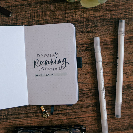 A dot grid notebook is lying on a dark surface. The page of the journal spread says Dakota's Running Journal. There are two brush pens on the table next to the notebook.